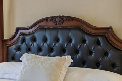 Close-up of a hand-carved solid wood headboard featuring intricate regency-style scrollwork and premium black tufted leather upholstery with nailhead trim.